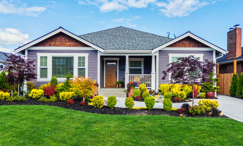 House with purple-blue siding on a partially cloudy day, with blue sky in the background. The foreground features shrubs, two planters with flowers, and a few trees.